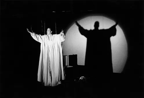 Schwarz-Weiß-Fotografie einer Person in einem langen Gewand auf einer Bühne, mit ausgebreiteten Armen vor einem kreisförmigen Lichtkegel und einem großflächigen Schatten an der Wand.