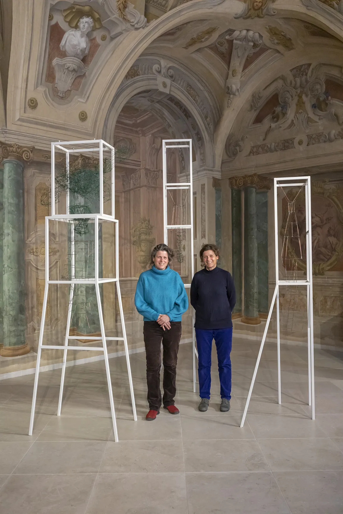 Zwei Fragen stehen neben Metall-Objekten in einem barocken Raum mit Fresken