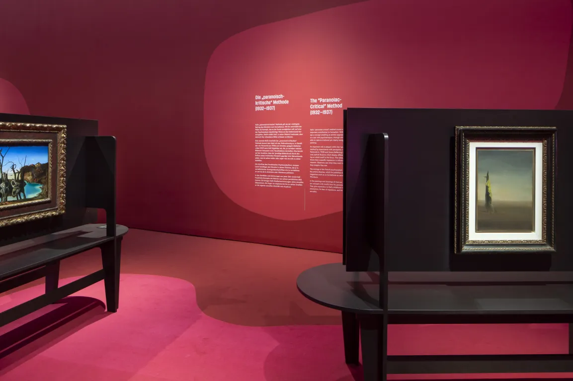 Ausstellungsraum mit roten Wänden, darauf surrealistische Bilder 