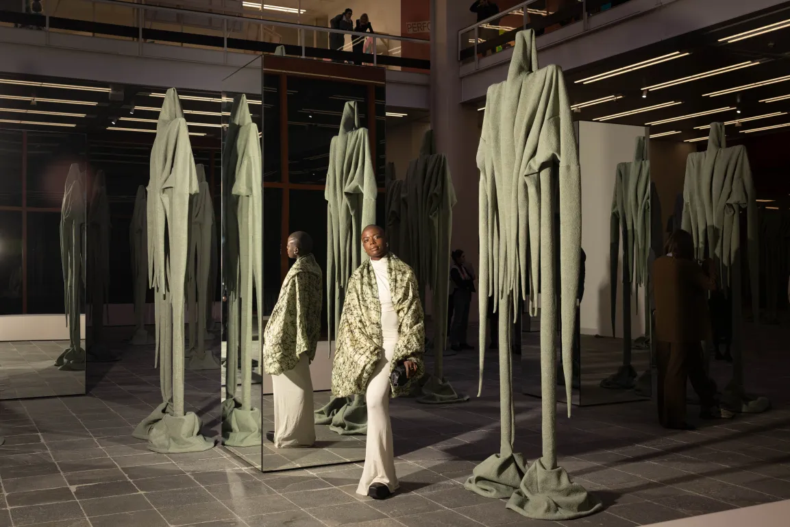 Die Künstlerin Sandra Mujinga steht inmitten ihrer Installation im Ausstellungsraum des Belvedere 21. Sie ist umgeben von überlebensgroßen drapierten grau-grünen Stoff-Figuren und Spiegeln.