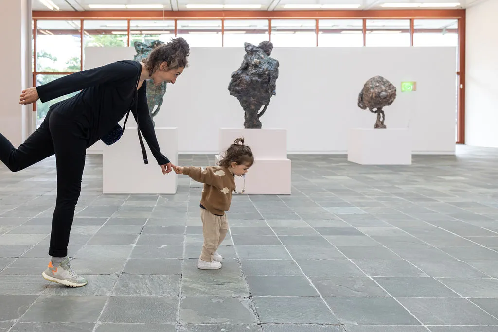 Ein Kleinkind führt bzw. zieht seine Mutter an der Hand durch den Ausstellungsraum des Belvedere 21. Im Hintergrund sind drei Skulpturen zu sehen.