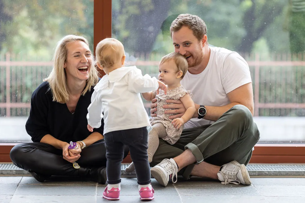 Eine blonde Frau und ein blonder Mann sitzen am Boden im Ausstellungsraum des Belvedere 21. Der Mann hat ein Kleinkind auf dem Schoß, während ein zweites auf die Gruppe zugeht.