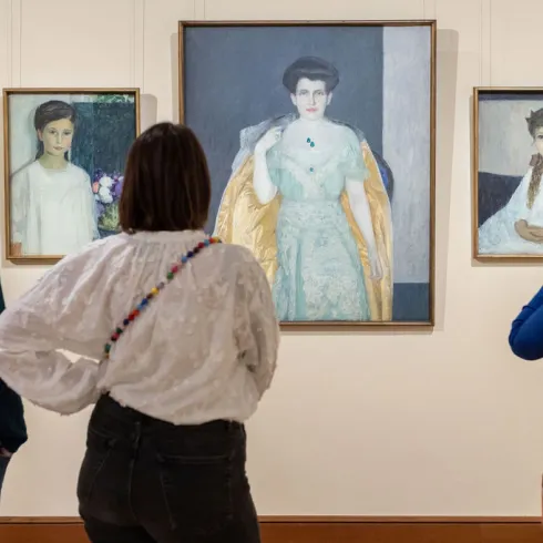 Drei Personen, von hinten gesehen, betrachten die Porträts einer Frau und zweier junger Mädchen.