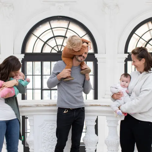 Von links nach rechts: eine Mutter mit Baby im Arm, ein Vater mit einem Kleinkind auf den Schultern und eine Mutter mit einem Baby auf der Schulter stehen vor einer weißen Balustrade und Fensterfront im Barockschloss.