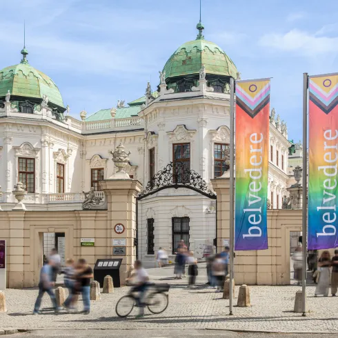 Ansicht des Belvedere-Park-Eingangstors auf der Prinz-Eugen-Straße. Im Vordergrund gehen Menschen , im Hintergrund sieht man das Obere Belvedere. Auf der rechten Bildseite wehen die Belvedere-Flaggen für den Pride-Month.