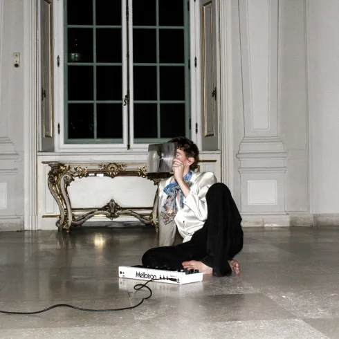 Oskar Haag sitzt barfuß auf dem Boden eines hellen Raums im Belvedere und hält sich ein Blatt Papier vor das Gesicht. Vor ihm liegt ein kleines elektronisches Musikinstrument.