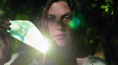 Filmstill Junge Frau mit dunklen Haaren die einen Spiegel neben ihrem Gesicht hält, der die Sonne reflektiert