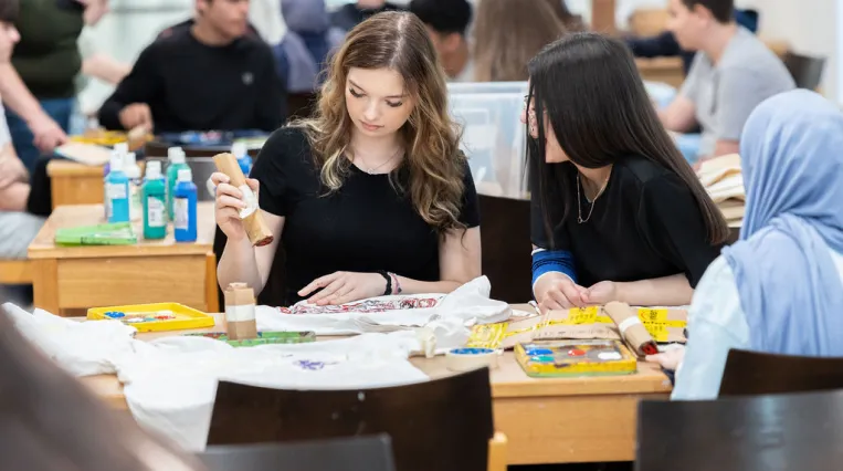 Jugendliche sitzen in einem Atelier und arbeiten mit Stempel aus Karton und Farbe an einem T-Shirt. Zwei Jugendliche sind im Fokus die restlichen Menschen sind unscharf im Hintergrund